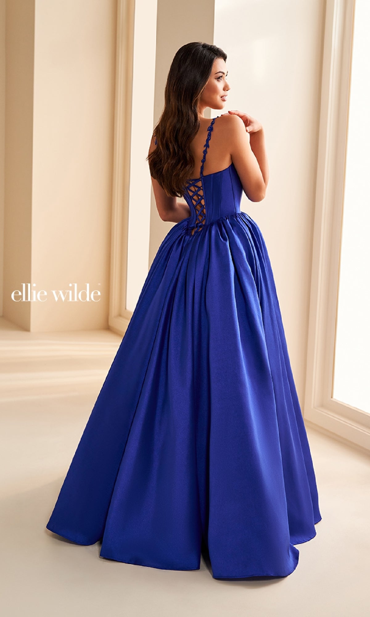 A woman in the Ellie Wilde EW36221 long prom dress, featuring a royal blue hue, corset bodice, and lace-up back, stands indoors near large windows.